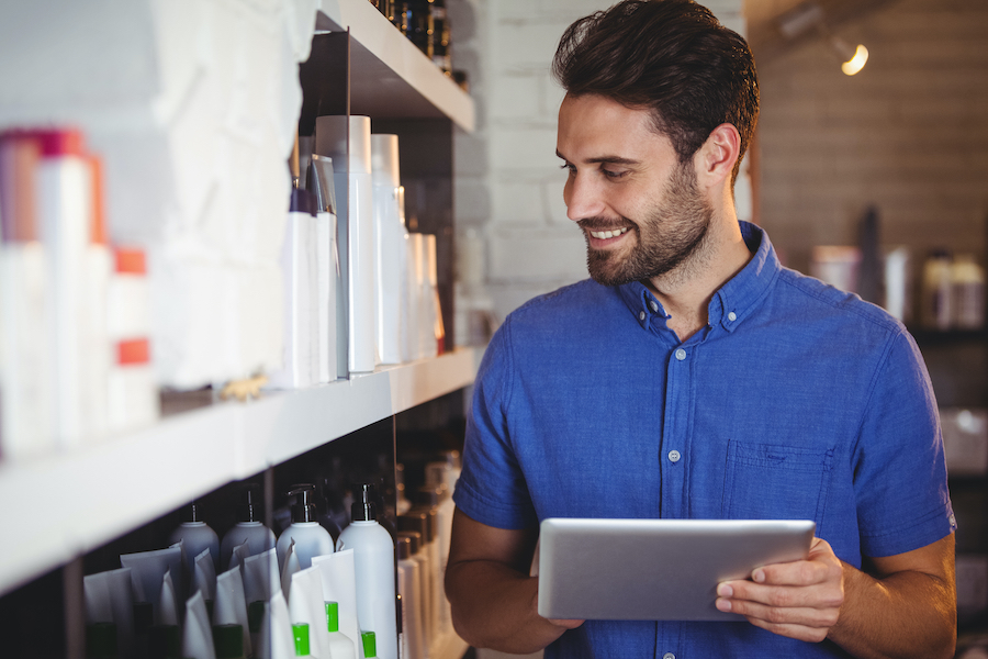 Ein lächelnder Saloninhaber im blauen Hemd schaut neben Regalen mit Haar- und Beautyprodukten auf ein Tablet.