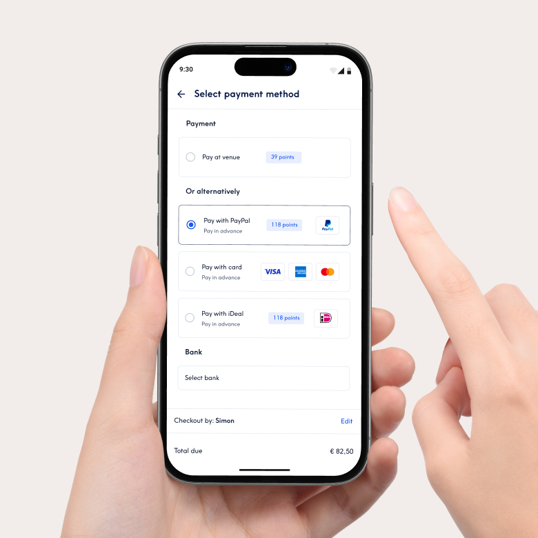Nahaufnahme eines Smartphones, das die Zahlungsoberfläche der Treatwell-App zur Auswahl einer Zahlungsmethode anzeigt. Zu den Optionen gehören die Zahlung vor Ort oder die Vorauszahlung mit PayPal, Karte oder iDeal. Eine Hand hält das Telefon und ein Finger zeigt auf den Bildschirm.
