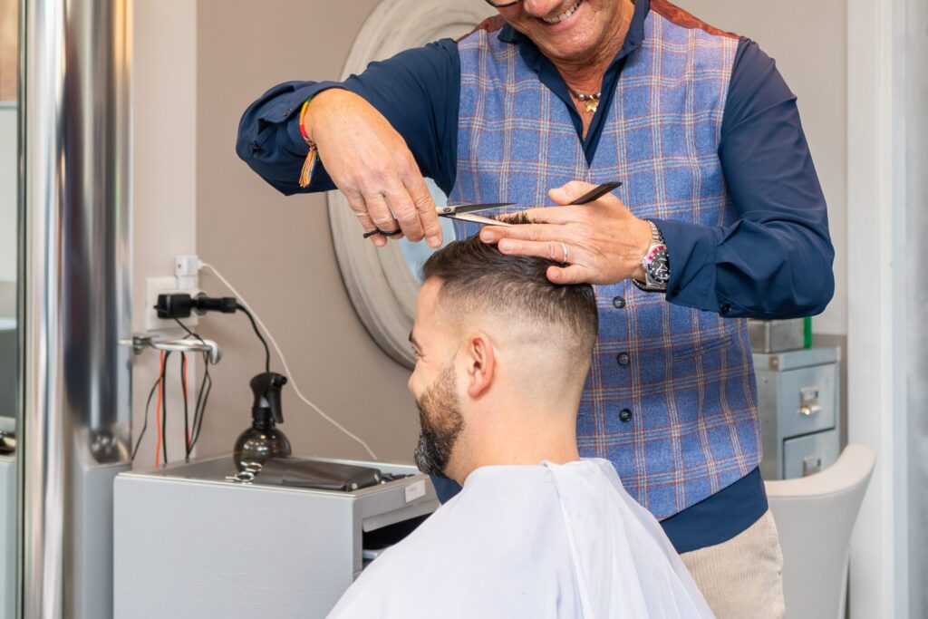 Ein Friseur in einer blau karierten Weste schneidet einem Kunden in einem Salon mit Schere und Kamm die Haare. Der Kunde hat kurze Haare, einen Bart und trägt einen weißen Friseurumhang.