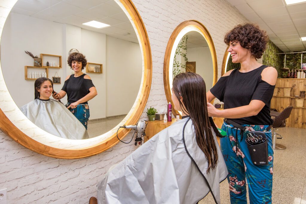 Eine lächelnde Friseurin mit kurzen lockigen Haaren bürstet das lange, feuchte Haar einer Kundin vor einem großen, runden, beleuchteten Spiegel. Die Szene spiegelt sich im Spiegel wider und zeigt das Innere des Salons.