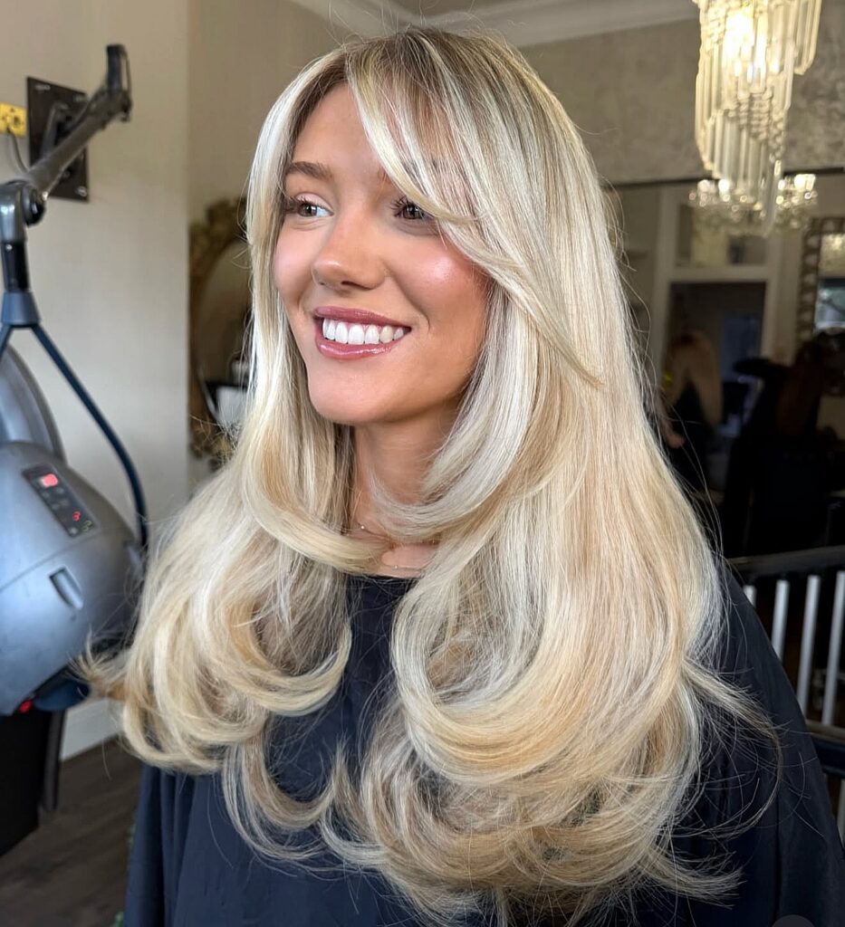 A smiling woman with long blonde hair styled into U-bangs that are shorter in the center and lengthen toward the ears, blending into soft face-framing layers.