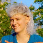 Eine junge Frau mit lockigem, weiblondem Vokuhila-Haarschnitt, die an einem sonnigen Tag drauen sitzt.