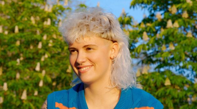 Eine junge Frau mit lockigem, weiblondem Vokuhila-Haarschnitt, die an einem sonnigen Tag drauen sitzt.