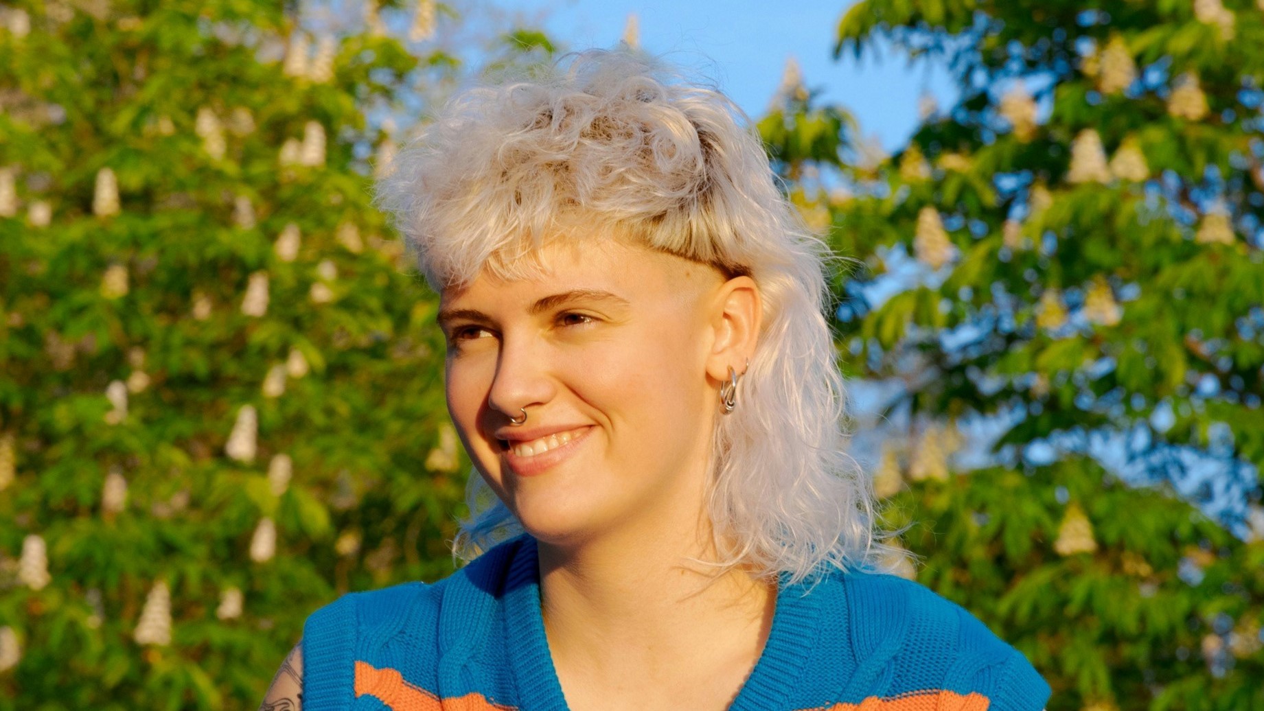 Eine junge Frau mit lockigem, weiblondem Vokuhila-Haarschnitt, die an einem sonnigen Tag drauen sitzt.