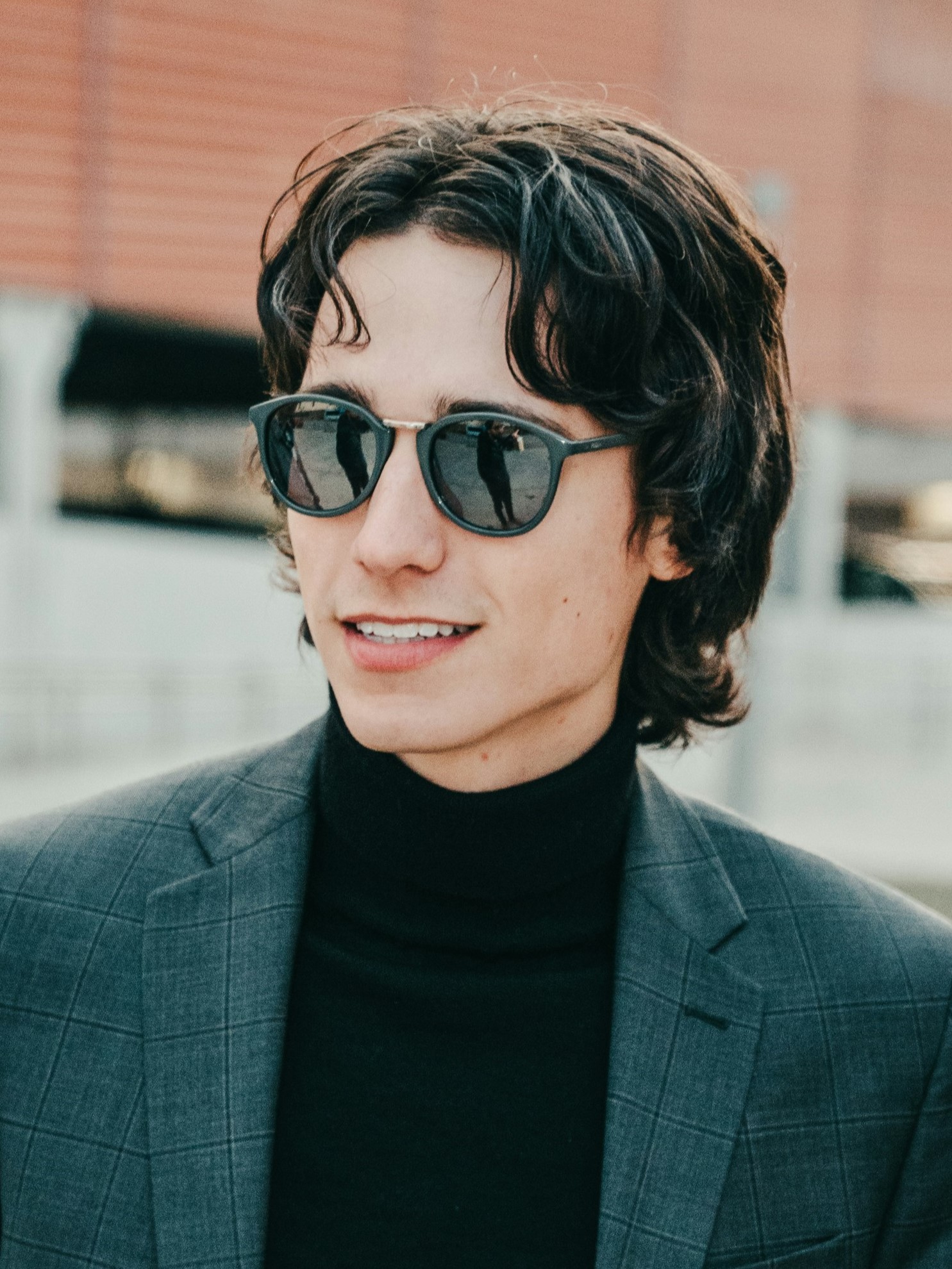 Ein junger Mann mit Retro-Vokuhila, der ein graues Sakko und Sonnenbrille trgt und drauen lchelnd steht.