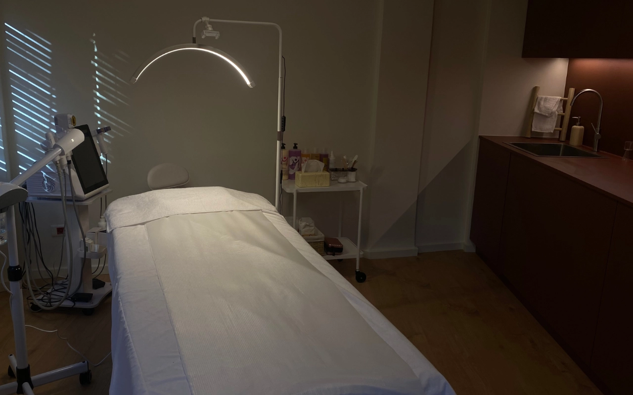 Ein konzentrierter Behandlungsraum mit weißem Bett unter einer Bogen-LED-Lampe, professioneller Ästhetik-Ausrüstung mit digitalem Bildschirm, Hautpflegeprodukten auf einem Trolley und dunklen Holzschränken mit Waschbecken an der hinteren Wand.