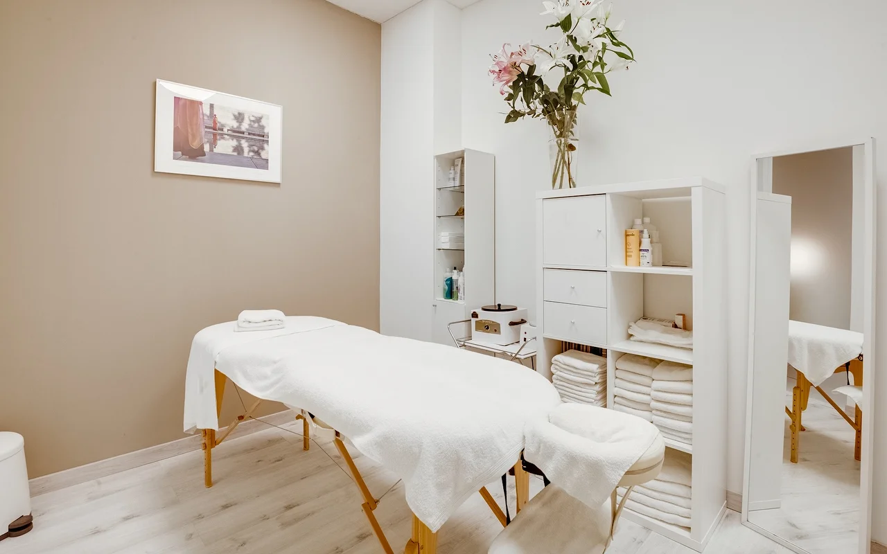 Ein ruhiger Behandlungsraum mit einer weien Massageliege und frischen Handtüchern. Der Raum hat neutrale Beige- und Weitöne, einen weien Schrank mit gefalteten Handtüchern und eine hohe Glasvase mit weien und rosafarbenen Lilien.