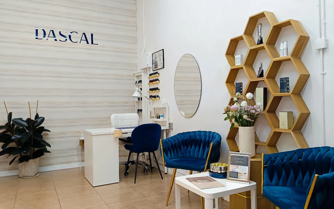 Ein stilvolles Beauty-Salon-Interieur mit tiefblauen Samtsesseln und einem sechseckigen Holzregal an der Wand. Ein weier Maniküretisch steht in der Ecke an einer hellen, texturierten Wand mit dem Schriftzug DASCAL in blauen Buchstaben.
