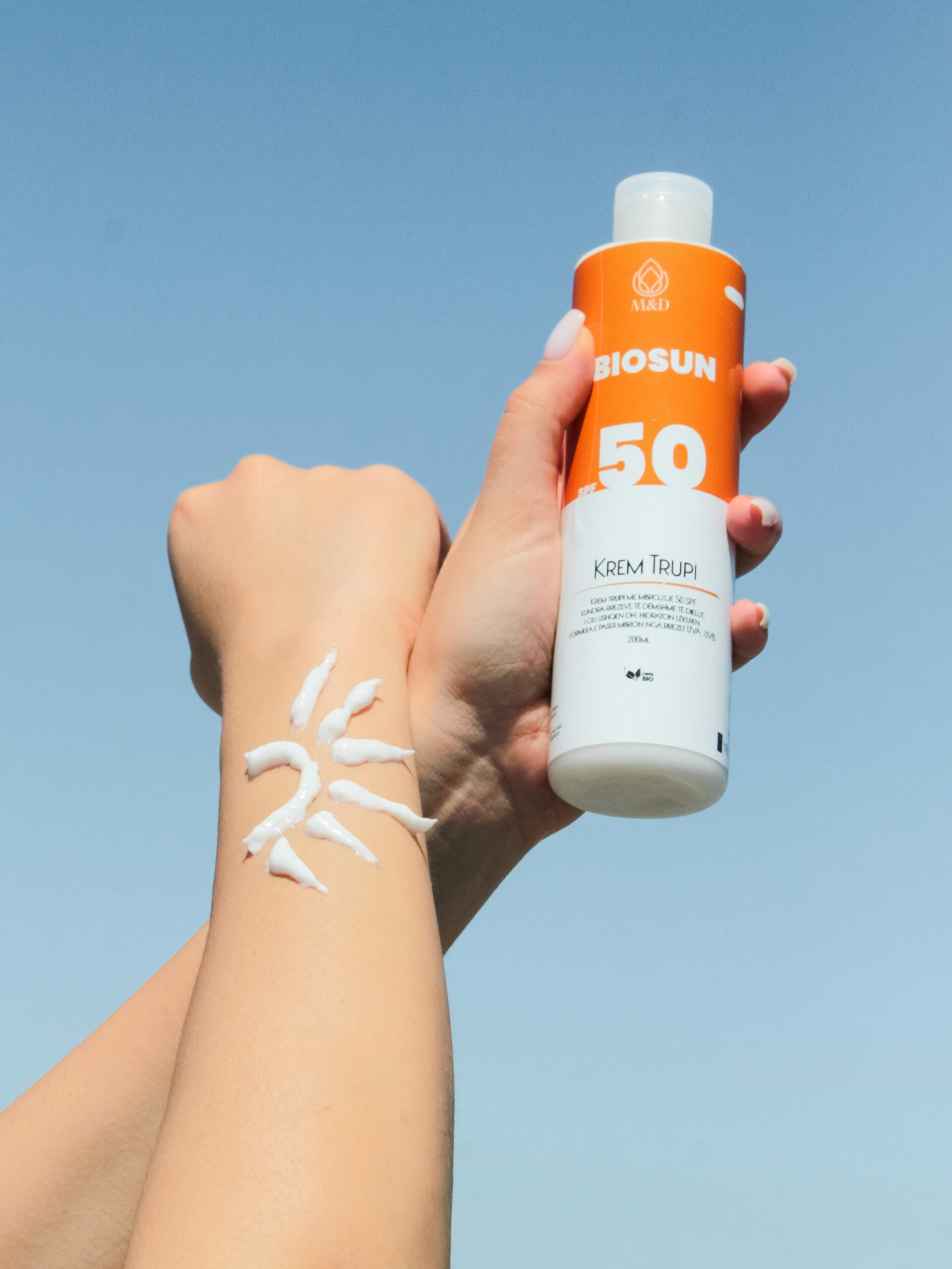 Nahaufnahme der Arme einer Frau vor blauem Himmel, eine Hand hält eine Flasche Sonnencreme und auf einem Unterarm ist Sonnencreme in Sonnenform aufgetragen