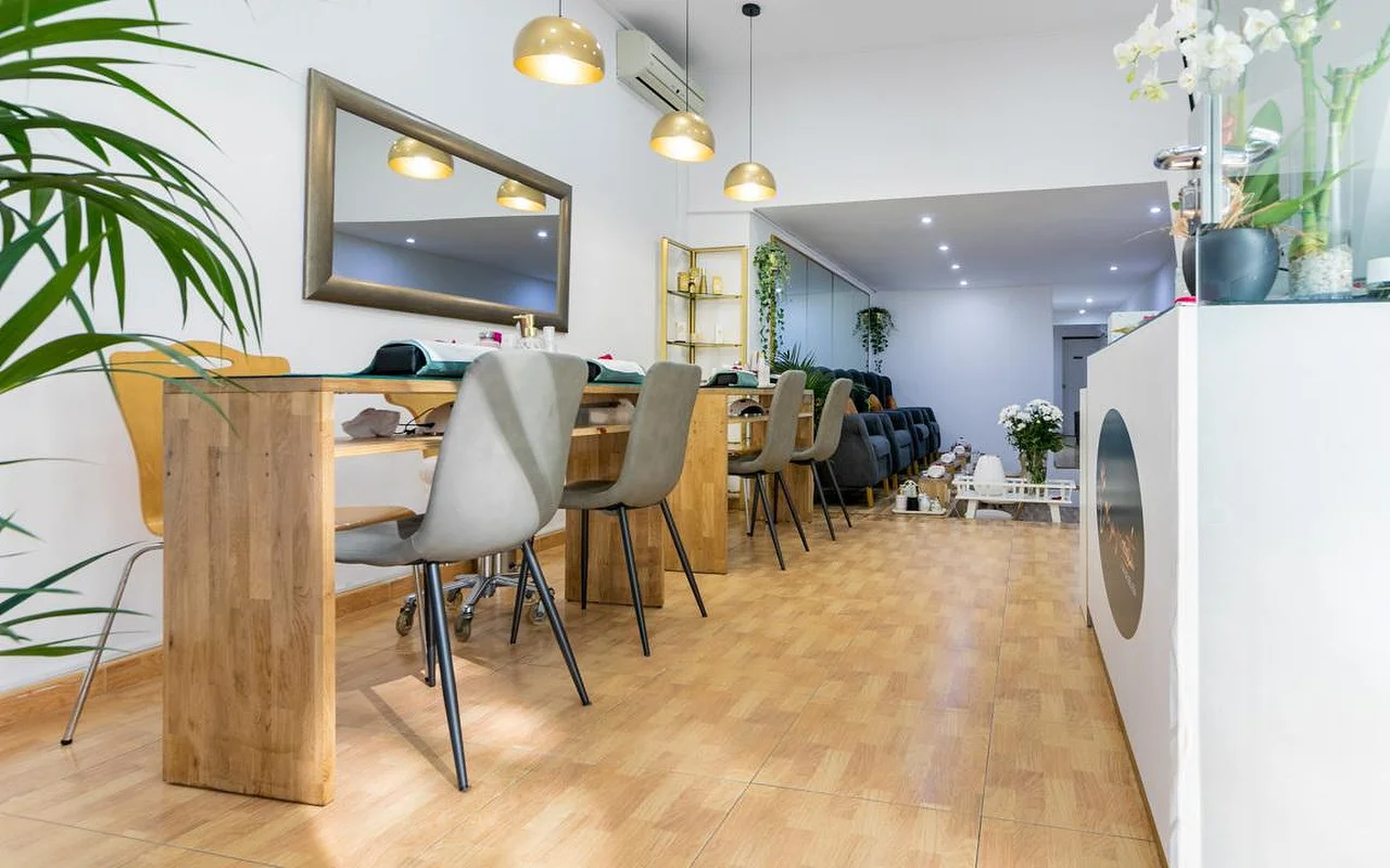Ein helles Nagelstudio mit langen Holz-Maniküretischen und grauen modernen Stühlen. Goldene Kuppel-Pendelleuchten hngen über den Pltzen, und ein groer Spiegel mit dunklem Rahmen hngt an der weien Wand.