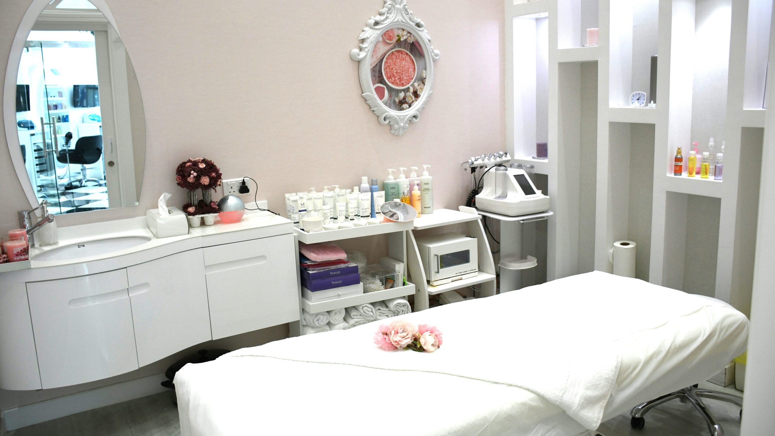 Heller, sauberer und einladender Behandlungsraum im Salon mit Liege, Produkten im Regal, einem verzierten Spiegel und Blumen