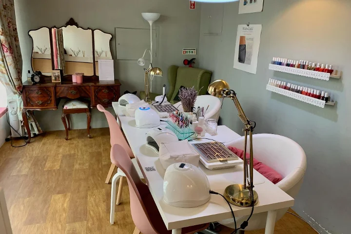 Innenansicht eines gemütlichen Beauty-Salons für Maniküre. Im Vordergrund ein langer weißer Tisch mit Nagellampen, Schreibtischleuchten und Werkzeugen, flankiert von rosa und weißen Stühlen. Im Hintergrund ein Vintage-Frisiertisch mit Dreifachspiegel, ein grüner Sessel und weiße Regale mit bunten Nagellackflaschen.