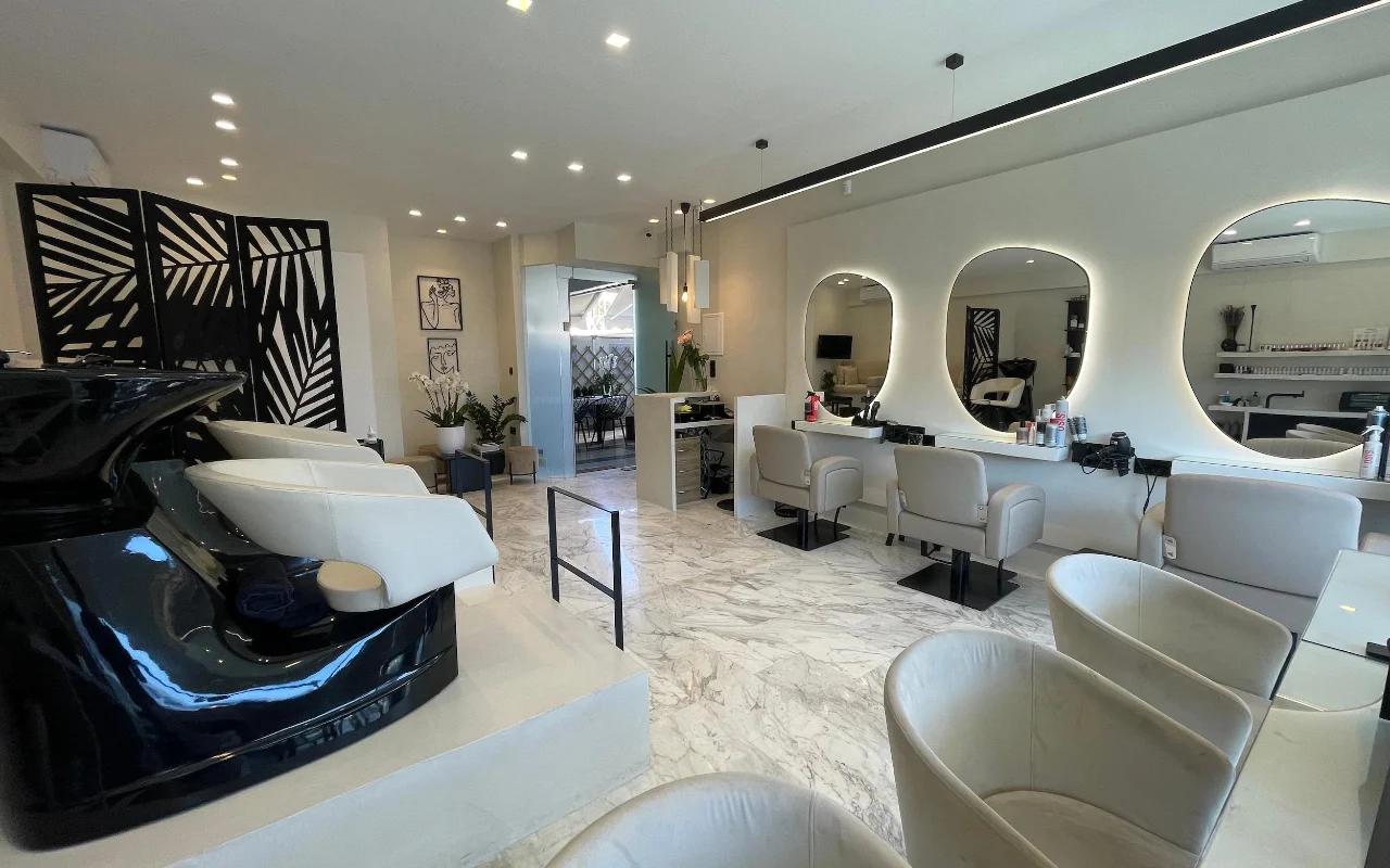 Ein eleganter Friseursalon mit weißem Marmorboden, cremefarbenen Leder-Stylingstühlen unter LED-gerahmten Ovalspiegeln, einer glänzenden schwarzen Waschstation und einem geometrischen Palmblatt-Raumteiler.