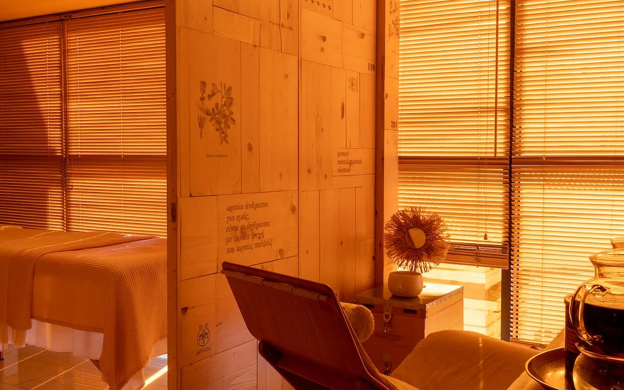 Ein warmer, bernsteinfarbener Spa-Behandlungsraum mit Holzvertäfelung, botanischen Illustrationen und griechischem Schriftzug an der Wand, einem Behandlungsbett mit Waffeldecke und dem APIVITA-Logo in der Ecke.