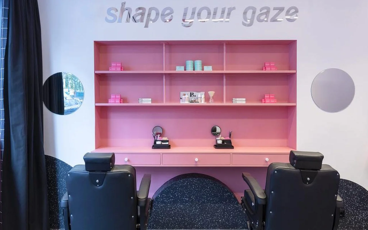 Ein spezialisiertes Brauen- und Wimpernstudio mit einem auffälligen pinkfarbenen Regal voller Beauty-Produkte, silbernem „shape your gaze“-Schriftzug an der Wand und schwarzen Leder-Behandlungsstühlen auf dunklem, gesprenkeltem Boden.