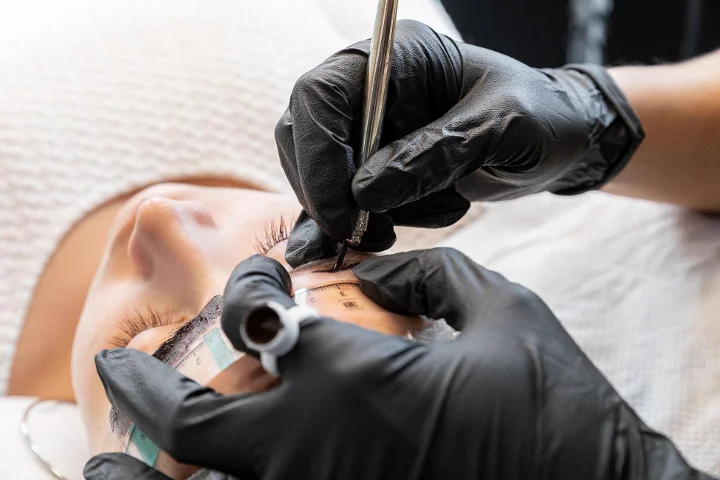 Nahaufnahme einer laufenden Augenbrauenbehandlung: eine Fachkraft mit schwarzen Handschuhen vermisst und markiert die Braue einer liegenden Kundin mit einem Messwerkzeug und einem Stift, die Augen geschlossen.