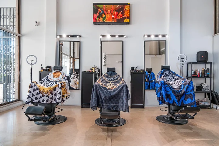 Ein heller Barbershop mit hohen Decken und drei schwarzen Barberstühlen in gemusterten Umhängen, jeder vor einem mit Glühbirnen gerahmten Spiegel, mit Ringlichtern, einem Wandbildschirm und hohen Fenstern.