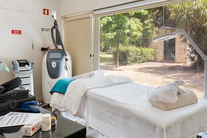 Ein Behandlungsraum mit einer weiß bezogenen Liege neben einem bodentiefen Fenster zu einem grünen Garten, einem Körperbehandlungsgerät auf einem Ständer, einer Lupenlampe und einem Glastisch mit Produkten.