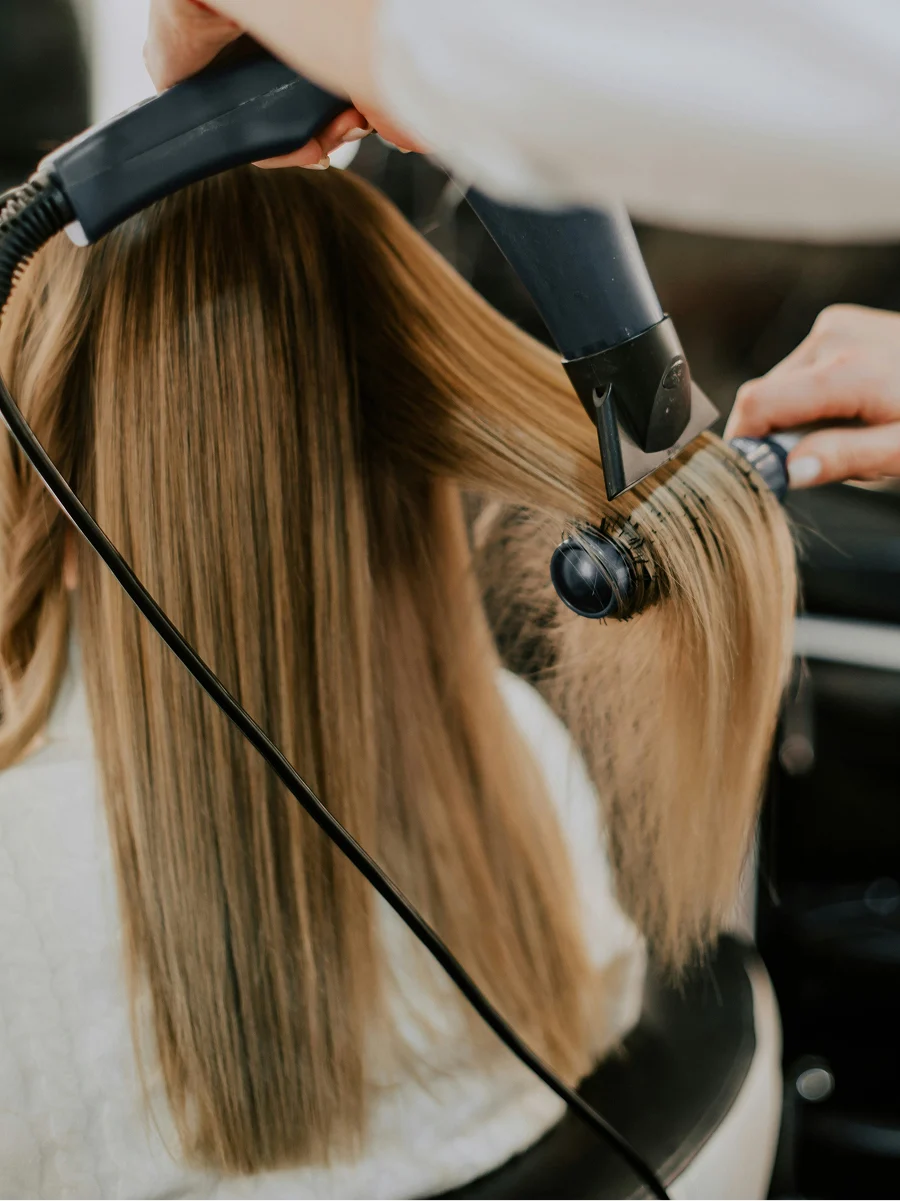 Rückansicht einer Frau beim Friseur, während eine Stylistin ihr Haar bei einem Keratin-Termin glatt föhnt
