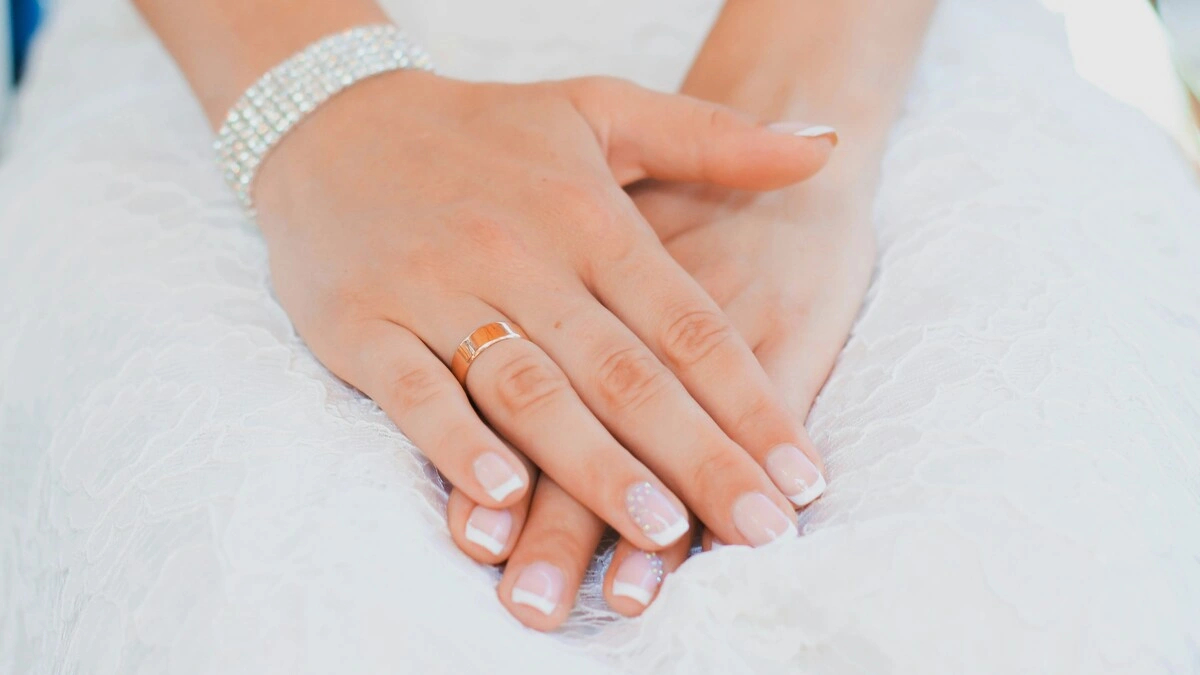 Die Hände einer Frau ruhen in ihrem Schoß; sie trägt ein Spitzen-Hochzeitskleid und eine gepflegte French-Tips-Maniküre in klassischer Kombination aus Nude und Weiß auf kurzen, eckig-ovalen Nägeln, mit zarter Verzierung an den Ringfingern