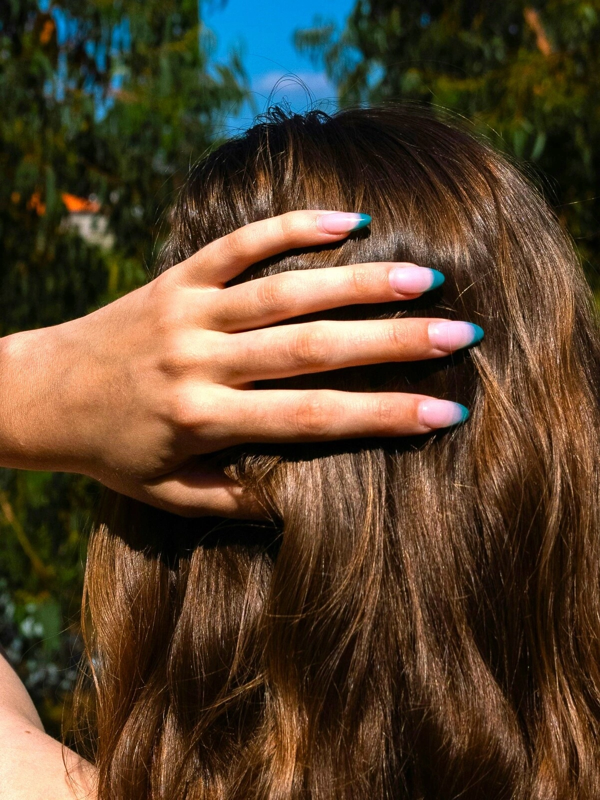 Die Hand einer Frau ruht an ihrem Hinterkopf und zeigt eine French-Tips-Maniküre mit zartrosa Basis und petrolfarbenen Spitzen auf mandelförmigen Nägeln