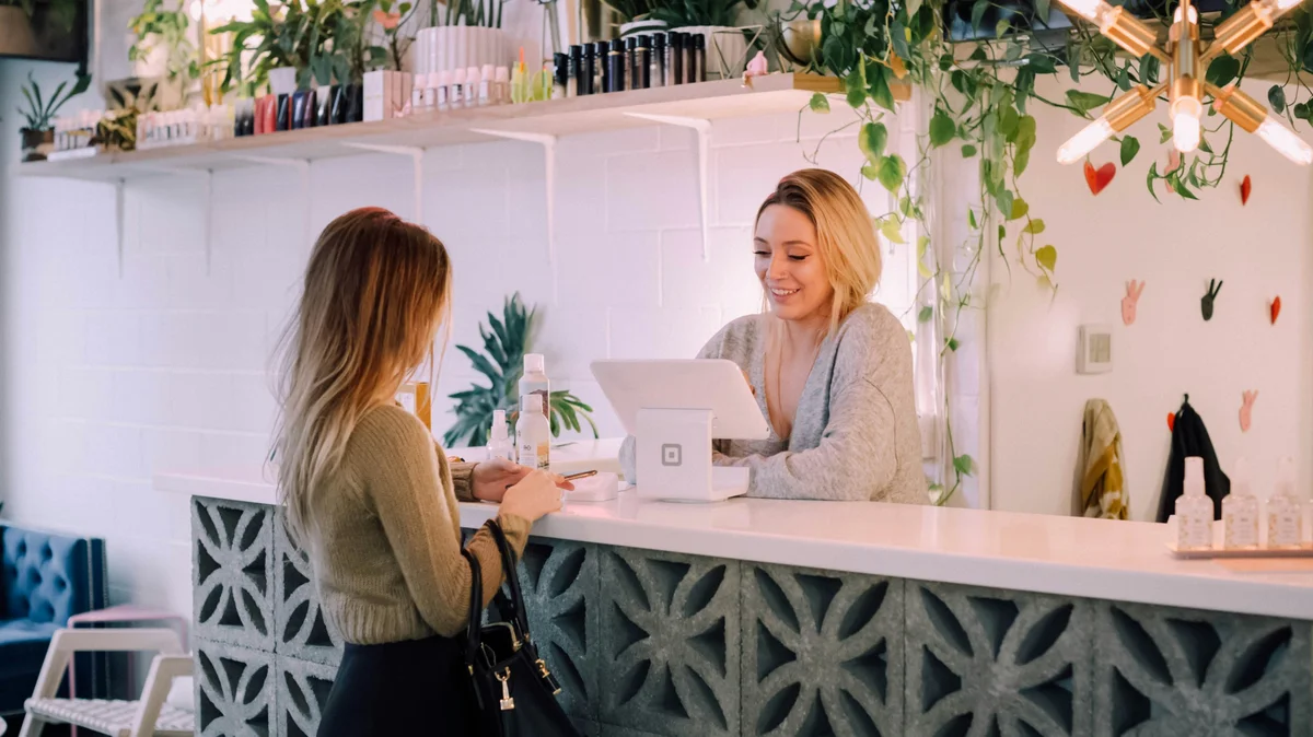 Eine Frau steht an einem Salontresen, während eine Rezeptionistin ihren nächsten Termin bucht; der Salon ist aufgeräumt, mit weißem Tresen, ordentlichen Produktregalen sowie Pflanzen und Blumen als Deko