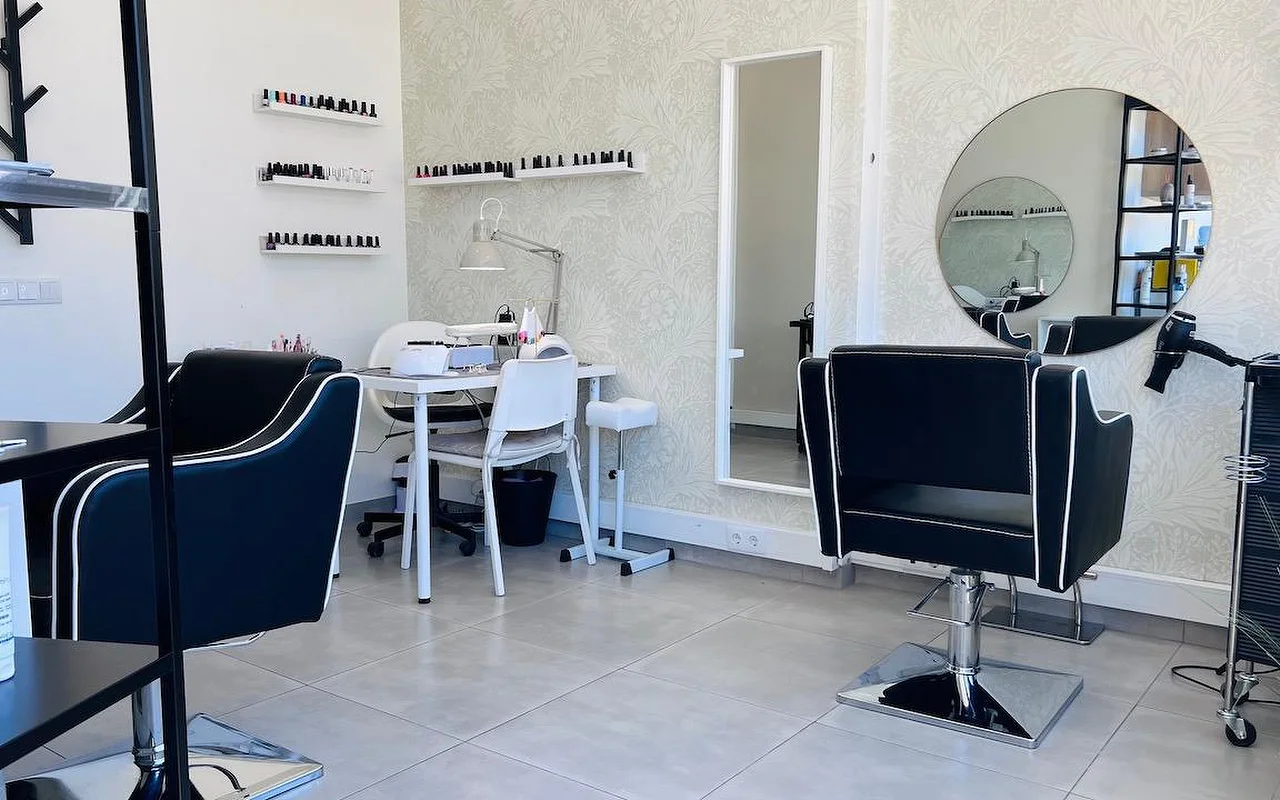 Ein kompakter Haar- und Nagelsalon mit schwarzen, weiß paspelierten Frisierstühlen, einem runden Spiegel und einem hohen Spiegel vor einer grauen Blümchentapete und Wandregalen voller Nagellacke.