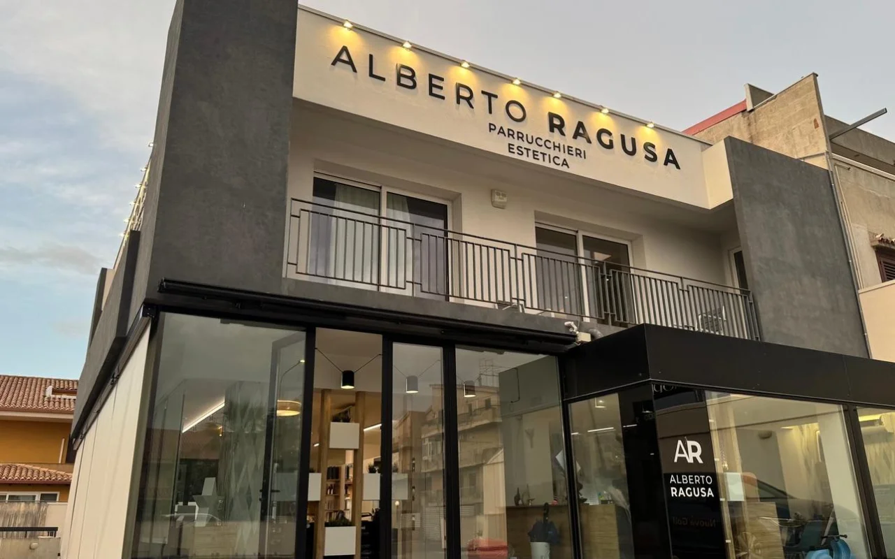 Das Äußere von Alberto Ragusa in der Dämmerung, ein zweistöckiger Salon mit einem großen beleuchteten Schild „Alberto Ragusa Parrucchieri Estetica“ über raumhohen Glasfenstern, mit warmem Licht, das von innen strahlt.