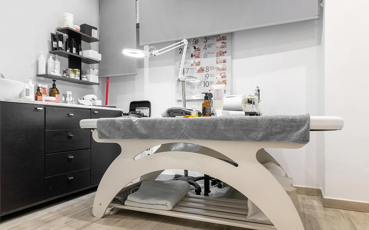 Ein kompakter Kosmetik-Behandlungsraum bei Atelier Beauty and the Lab, mit einer cremefarbenen Liege, einer verstellbaren Lupenlampe, dunklen Wandregalen voller Hautpflegeprodukte und einer nummerierten Behandlungsschritt-Tafel an der Wand.