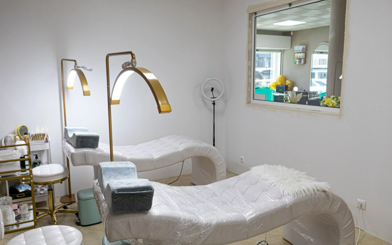 Ein heller Beauty-Behandlungsraum mit zwei weißen gesteppten Liegen, hohen goldenen Bogenlampen und einem Ringlicht, mit einem Innenfenster mit Blick zur Rezeption.