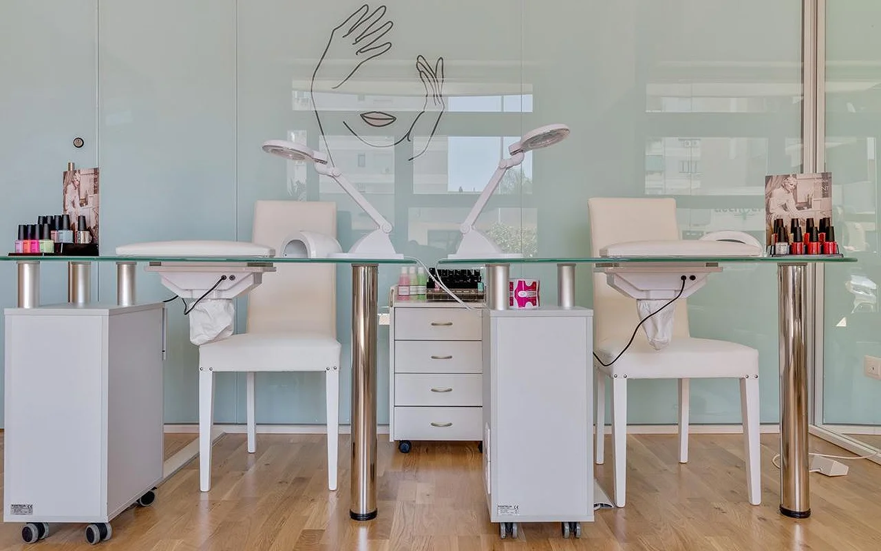 Ein Nagelstudio bei Perfetta Urban Beauty, mit zwei Maniküretischen mit Glasplatte und weißen Stühlen, Arbeitsleuchten, bunten Lack-Displays und einer mintgrünen Glaswand mit der Strichzeichnung eines Gesichts und zweier Hände.