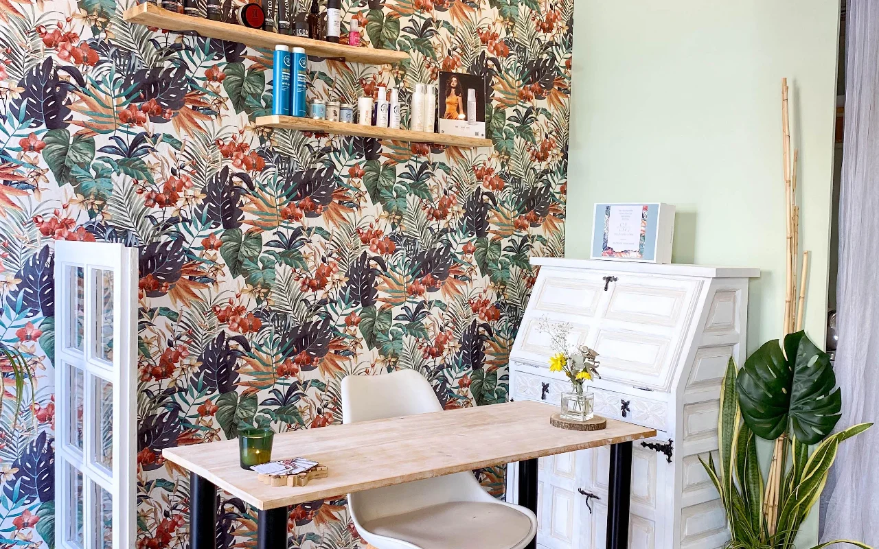 Ein Styling-Platz bei Shangara Lora Organics in Marbella, mit auffälliger tropischer Tapete, einem Holztisch, einem antiken weißen Sekretär und Bio-Haarpflege auf schwebenden Regalen.