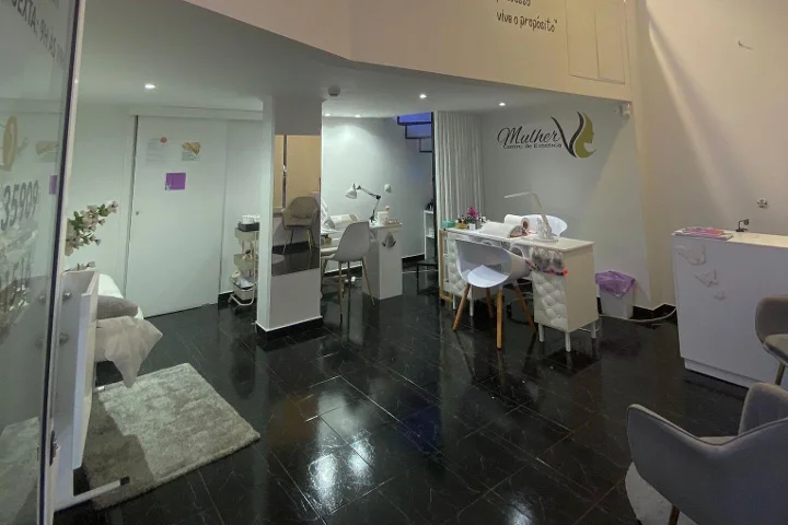 Innenraum eines Kosmetikstudios: dunkler polierter Fliesenboden, weißer Maniküre-Tisch und dezent beleuchtete Behandlungsbereiche, das Mulher-Logo an der Rückwand und eine Gesichtsbehandlungsliege hinter einer Glastrennwand sichtbar.