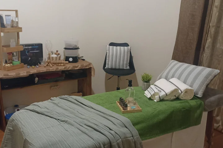 Gemütlicher Massageraum mit einer mit einem grünen Handtuch bezogenen Liege und aufgerollten weißen Handtüchern, einer Schale ätherischer Öle, Holz-Massagewerkzeugen auf einem rustikalen Beistelltisch und einem Hocker neben einer durch einen Vorhang abgetrennten Ecke.
