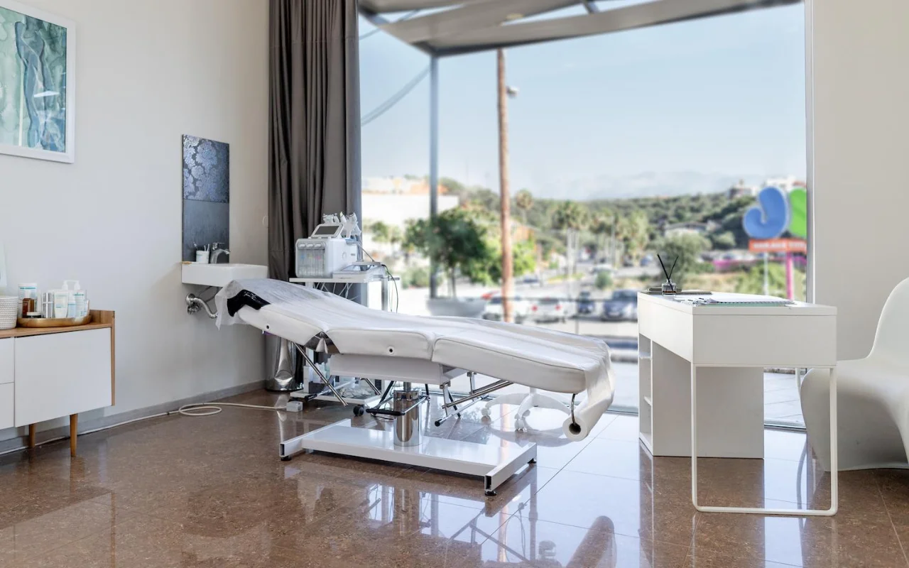 Ein sauberer weißer Behandlungsraum mit einer verstellbaren Beauty-Liege, Skincare-Geräten auf einem Wagen, gerahmten Kunstwerken und einem großen Fenster mit Blick auf Bäume, eine Straße und ferne Hügel.