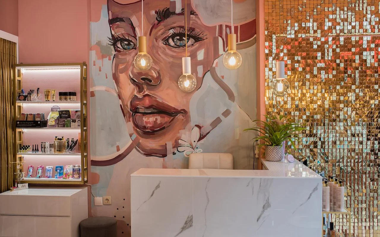 Ein Salon mit rosa Wänden mit einem großen gemalten Wandbild eines Frauengesichts, einer schimmernden goldenen Paillettenwand, einer Rezeptionstheke aus weißem Marmor und Pendelleuchten mit freiliegenden Glühbirnen.