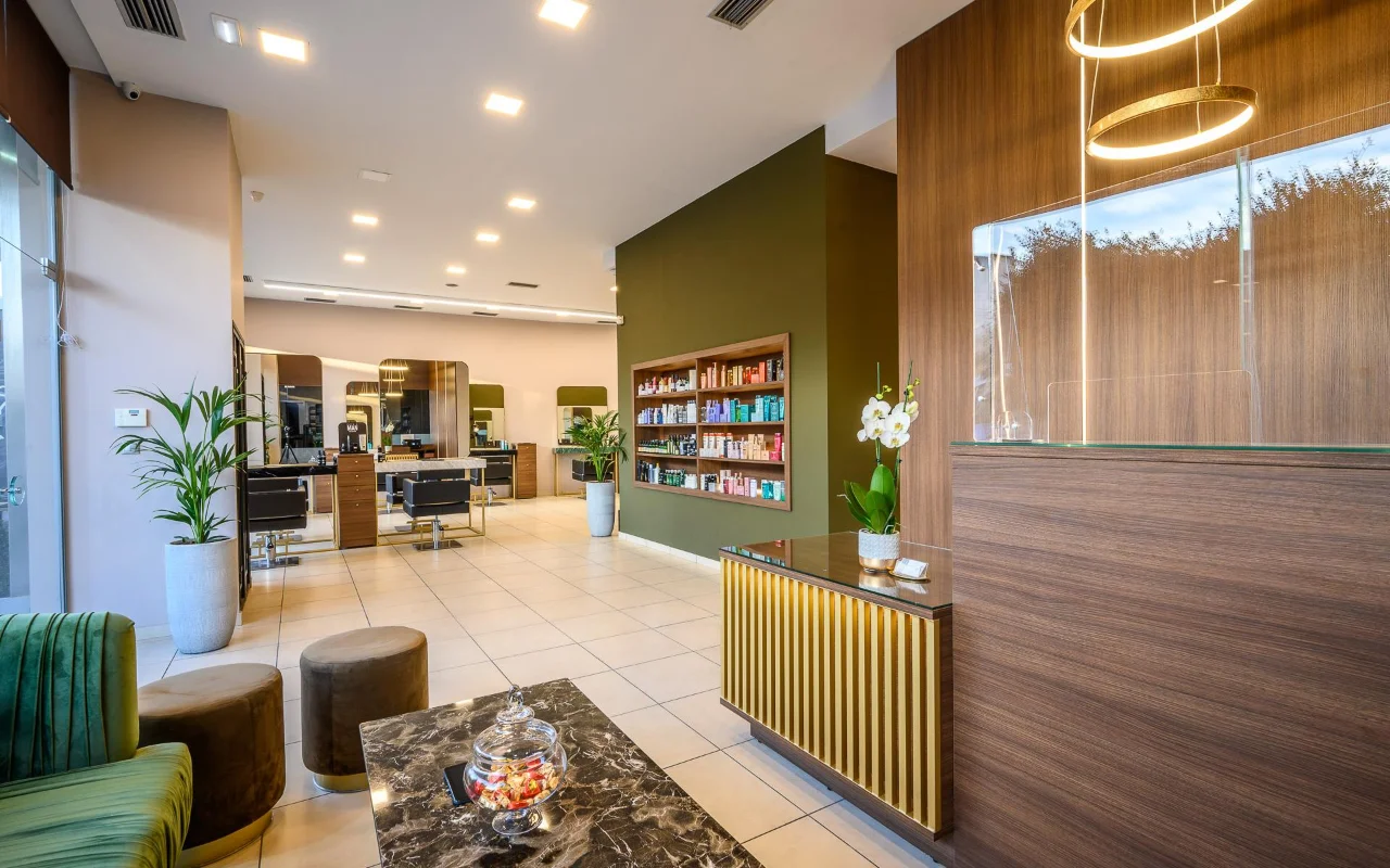 Ein gehobener Friseursalon mit warmen Holzvertäfelungen, einer olivgrünen Akzentwand aus Produkt-Regalen, Stylingplätzen mit großen Spiegeln, einer Rezeptionstheke mit goldenen Lamellen und grünen Samtsitzen unter kreisförmigen goldenen Pendelleuchten.