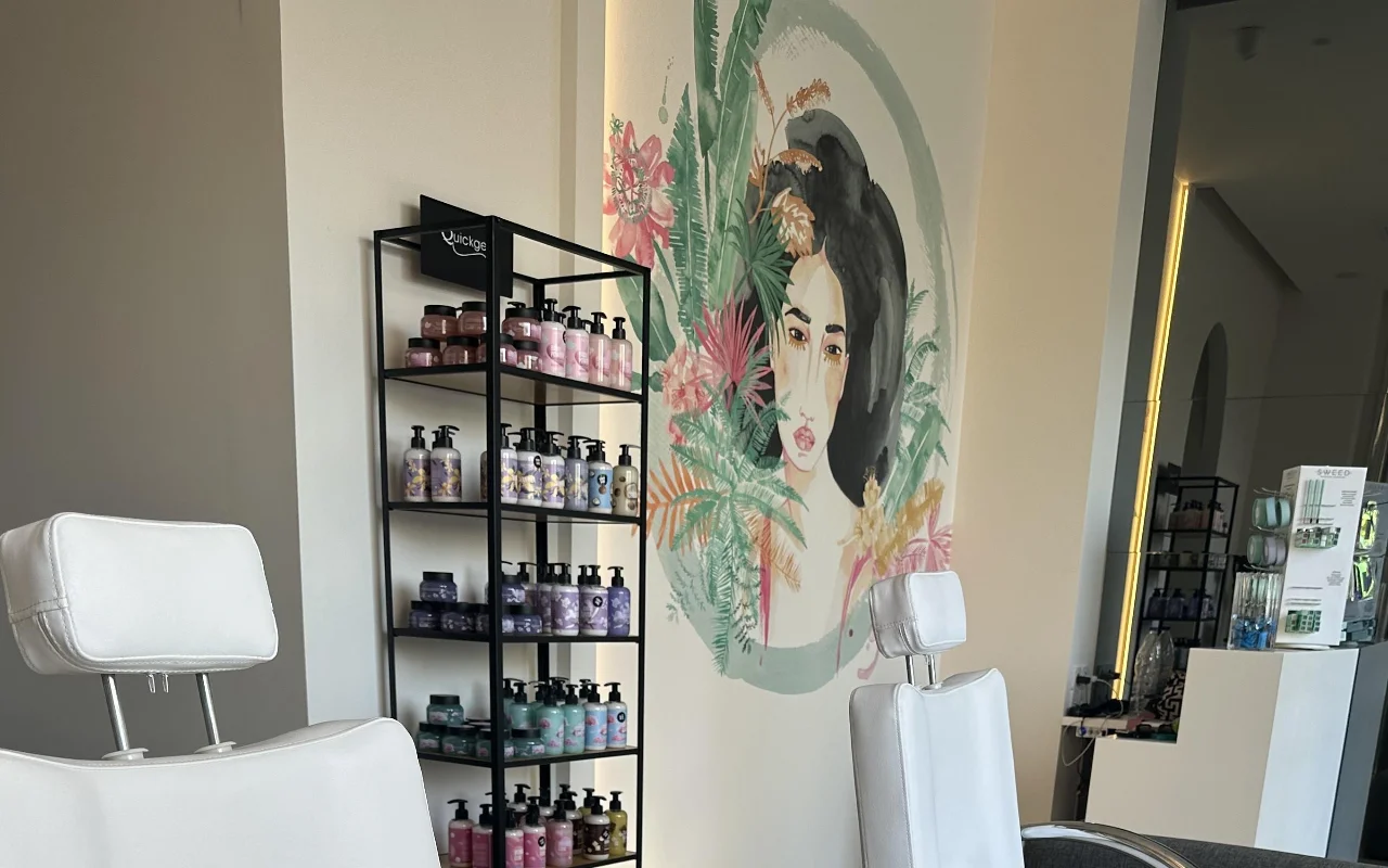 Eine Studio-Ecke mit zwei weißen verstellbaren Behandlungsstühlen, einem schwarzen Produkt-Regal, einem hintergrundbeleuchteten Spiegel und einem floralen Wandbild eines Frauengesichts, umrahmt von tropischem Laub.