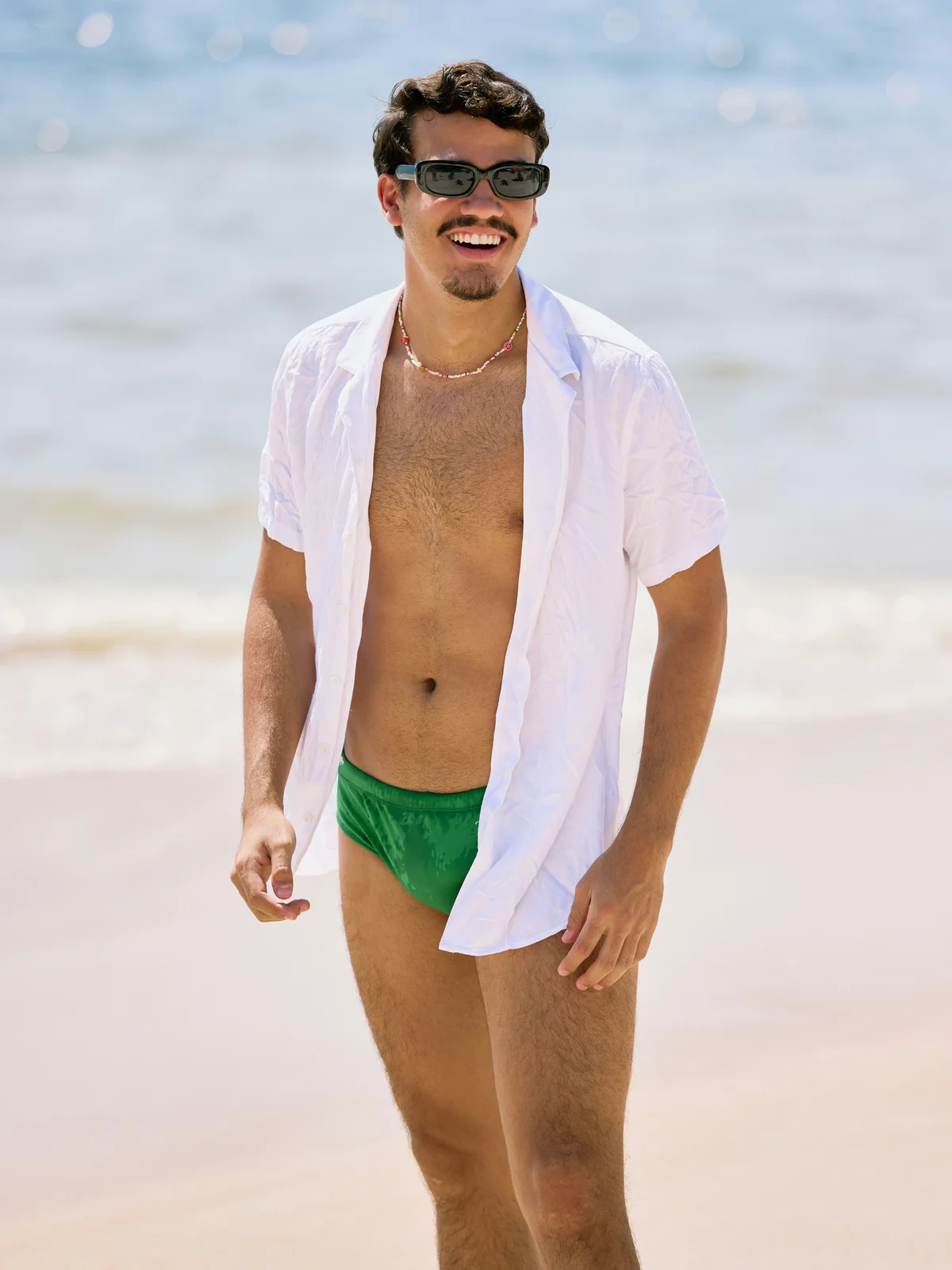 Ein lächelnder Mann steht an einem sonnigen Tag am Strand, er trägt eine grüne Badehose, ein offenes weißes Hemd und eine schwarze Sonnenbrille