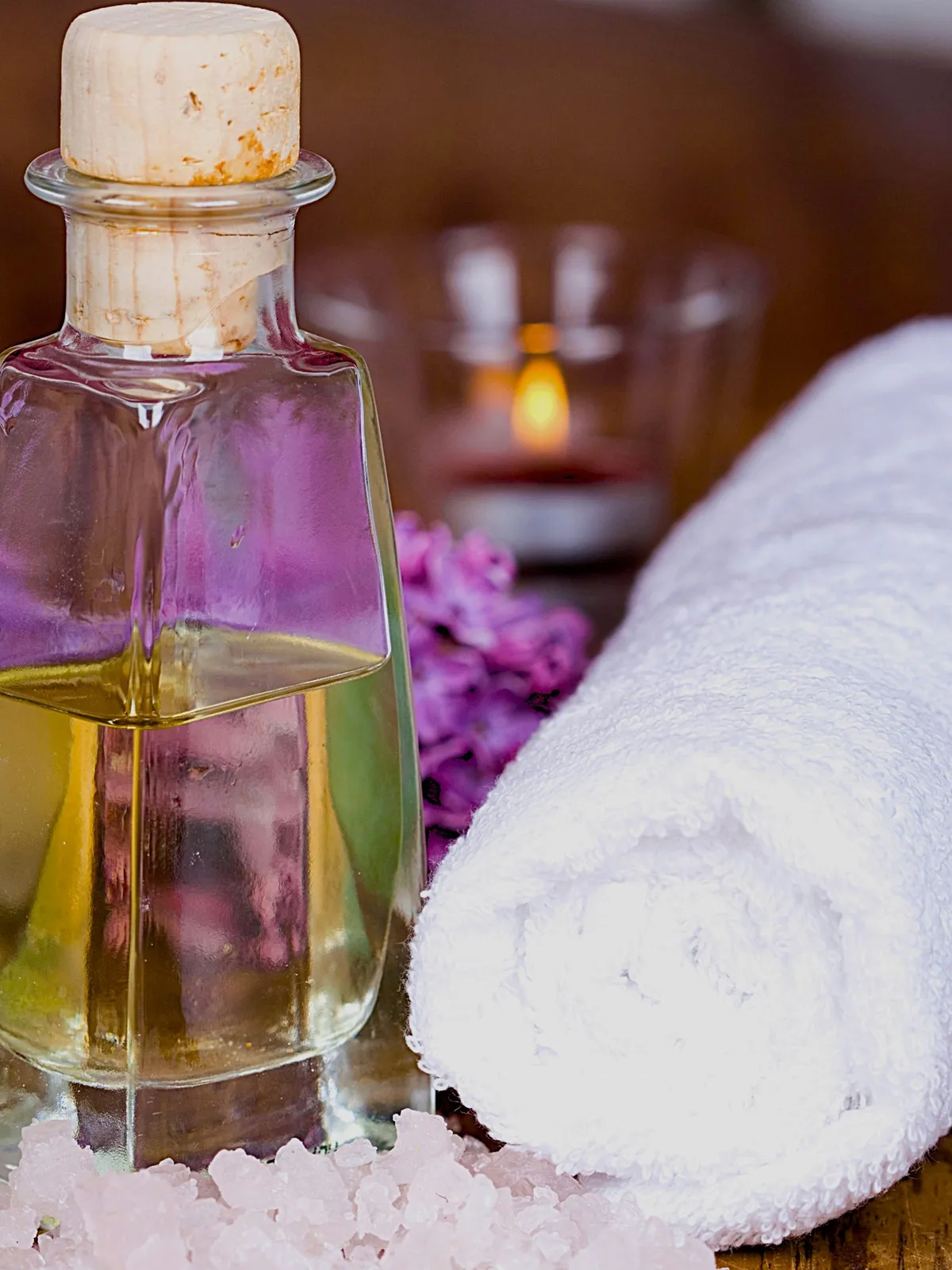 Nahaufnahme einer Glasflasche Öl mit Korken auf einem Tisch, daneben ein gerolltes weißes Handtuch und rosa Blüten